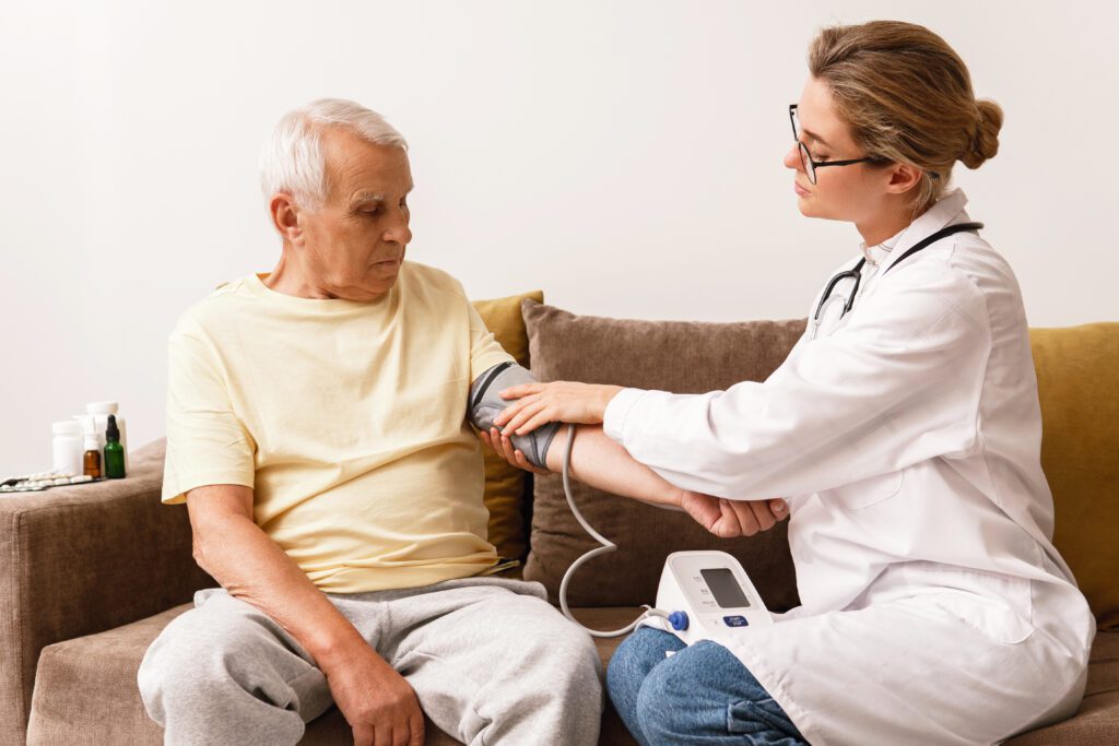 Doctor checks elderly man's blood pressure at home on a couch, with medical supplies on the table nearby.