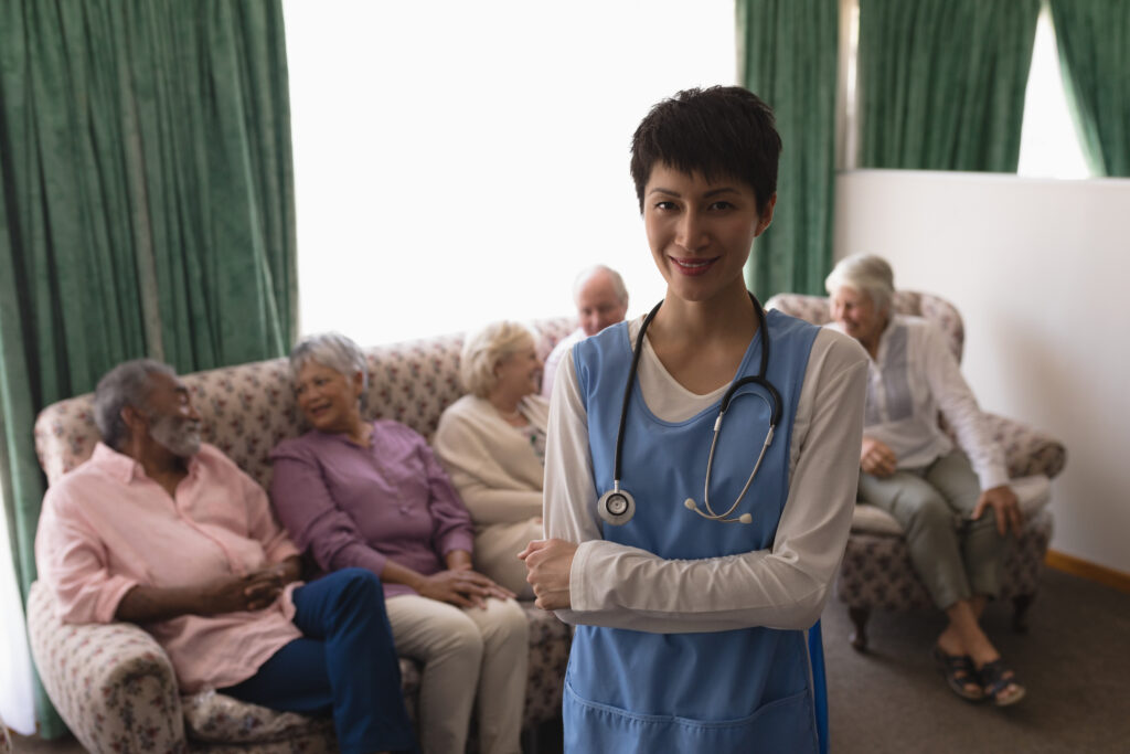 Front view of female doctor standing with arms crossed with senior person behind her on the sofa in living room at home